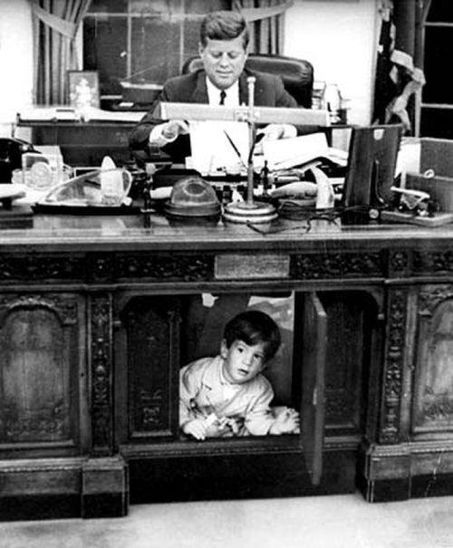President Kennedy, working late at his White House office, wears a slight smile on his face, indicating perhaps he is not completely unaware that his son, John Jr., is exploring under his desk in the Oval Office in the White House in 1963. John Jr. called the spot under the desk "my house" and was peeking from behind the "secret door." (AP Photo/Look Magazine, Stanley Tretick, File)
